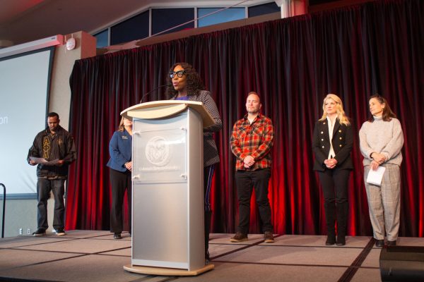 A woman stands at a podium and delivers a speech while five people stand behind her