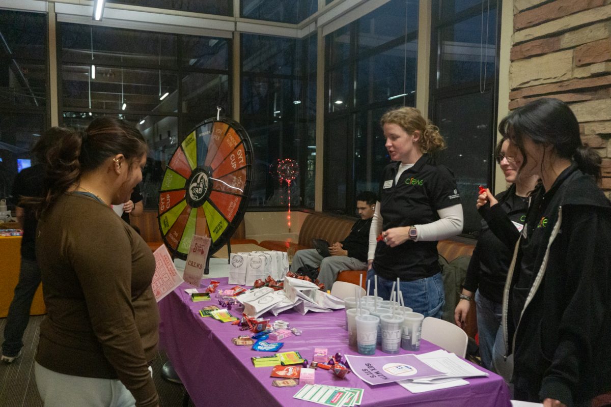 Three people stand behind a table with items on it talking to another person.