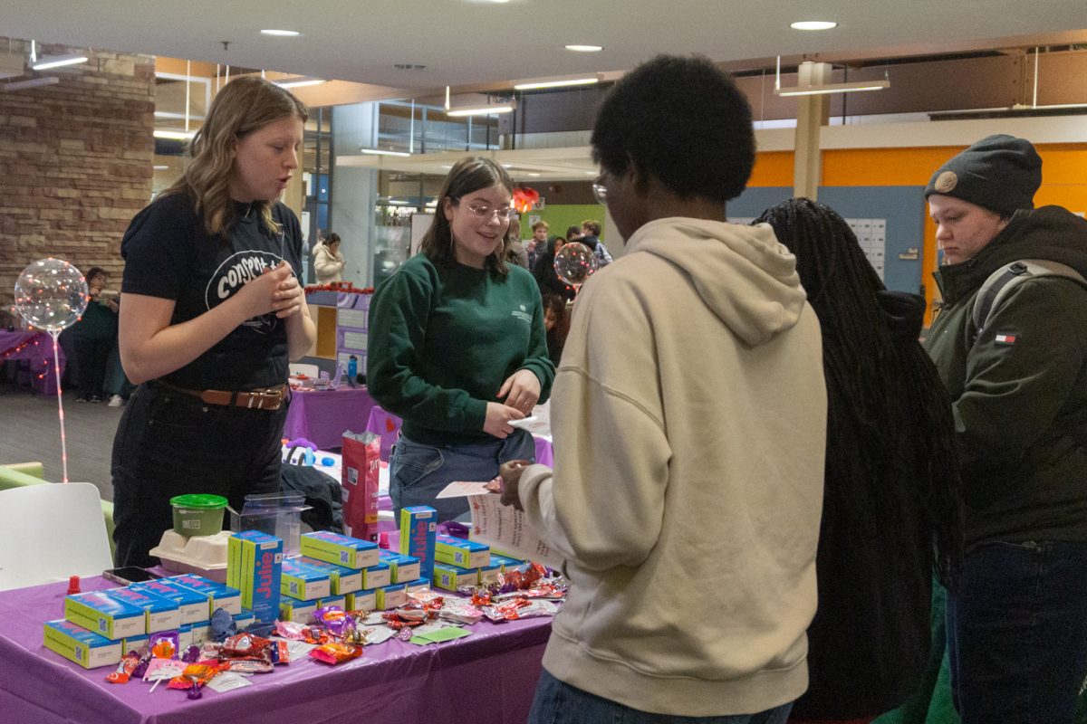 Two people stand behind a table with items on it, three people stand in front of the table looking at the first two.
