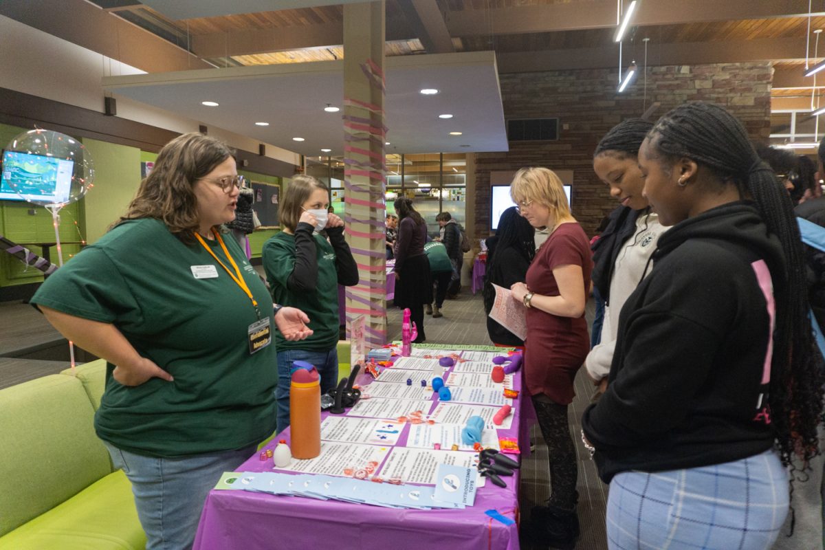 Two people stand behind a table with papers and sex toys on it while three people look at the table.