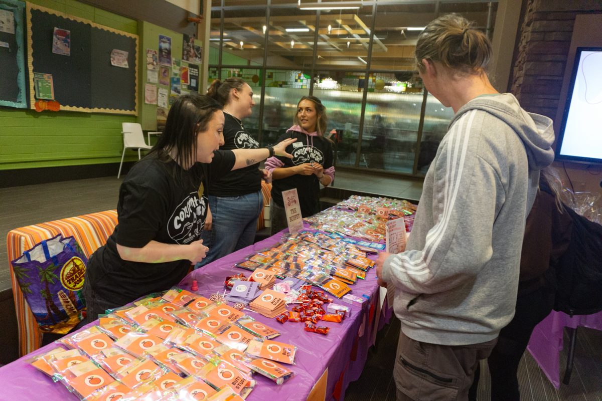 Three people stand behind a table, one is pointing at something on the table while another person listens.