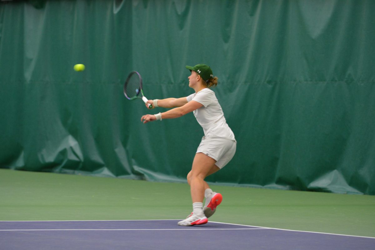 Girl in white tennis outfit and green hat hits the tennis ball.