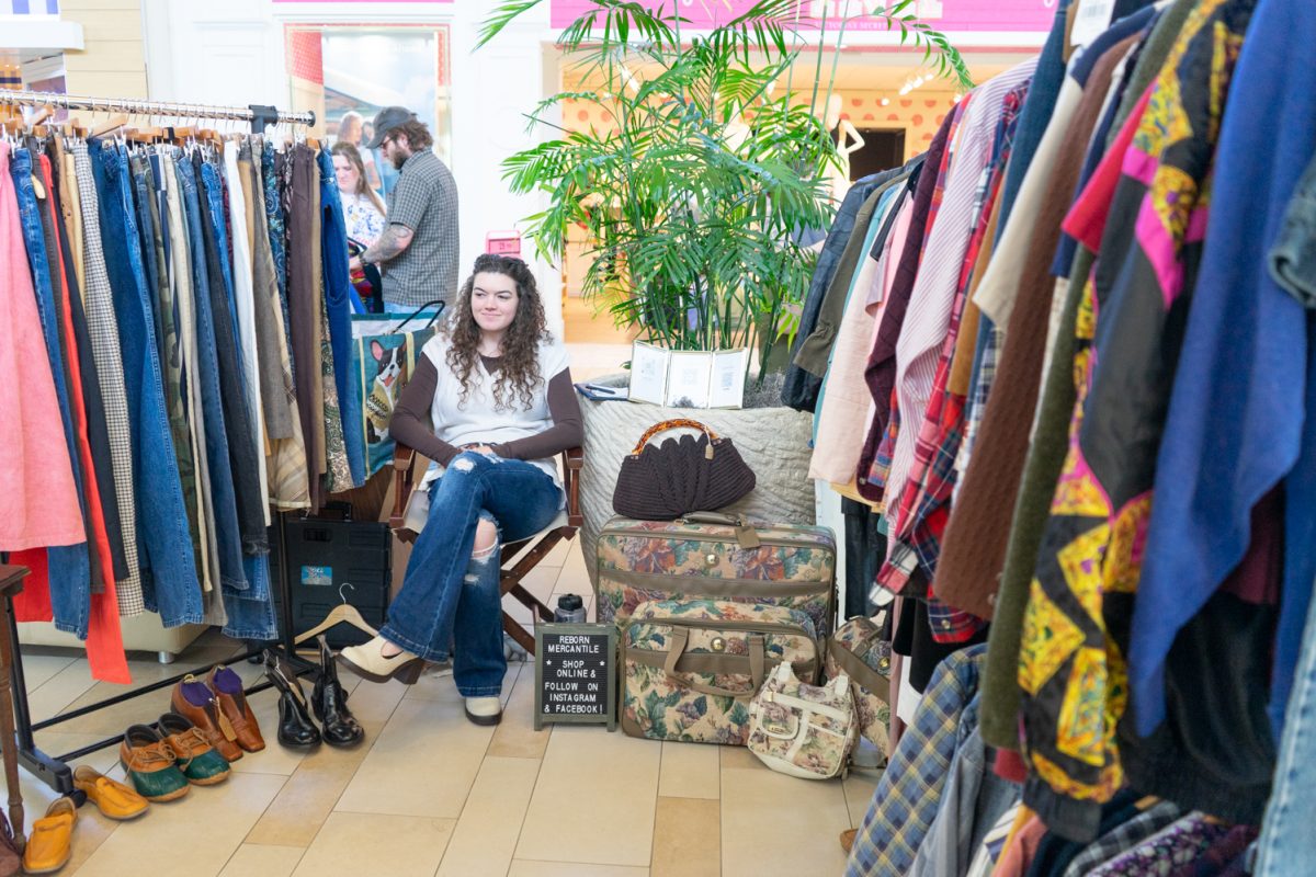 A woman with curly hair sits in a chair surrounded by vintage clothing and shoes.