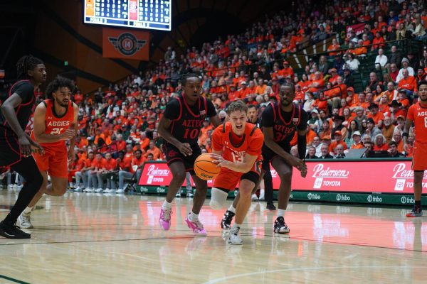 A player in orange falls while holding the basketball. Two defenders in black run behind him