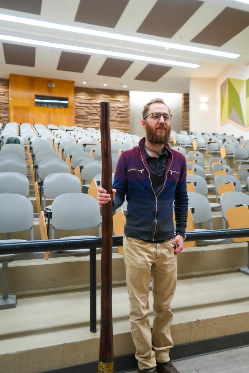 Dr. Robin Rothfeder stands in front of a lecture hall holding his didgeridoo which stands at about the same size as him.