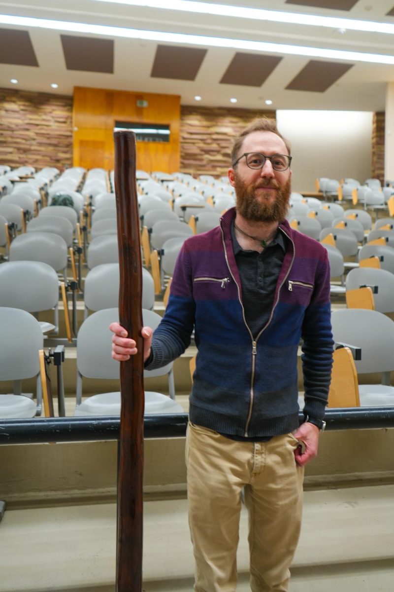 Dr. Robin Rothfeder stands in front of a lecture hall holding his didgeridoo which stands at about the same size as him.