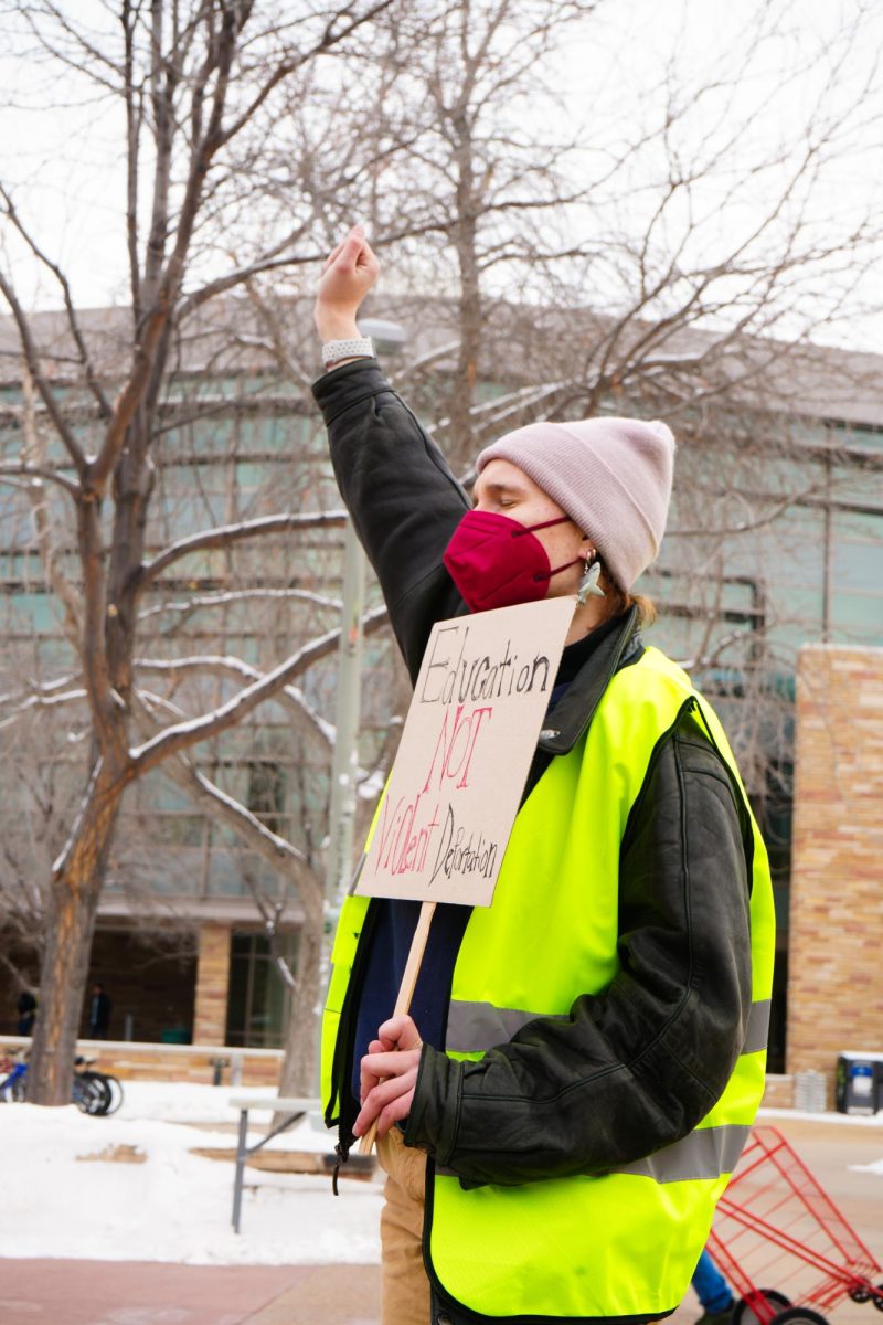 A person protesting in front of the Lory Student Center holds their fist up and holds a sign that reads, "Education, not Violent Deportation."