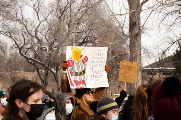 A person in a group holds up a sign, chanting. The sign reads, "Mirror, mirror, on the wall, who is the most racist, fascist of them all?" and features a caricature of President Donald Trump.