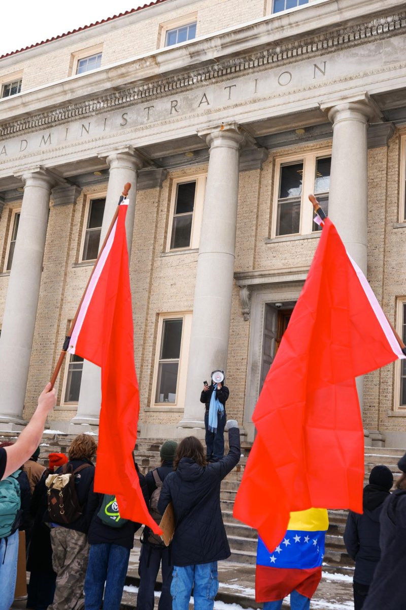 Two red flags wave in front of the CSU administration building in the crowd of protesters. In the middle of the flags, a person up on the steps addresses the crowd through a megaphone.