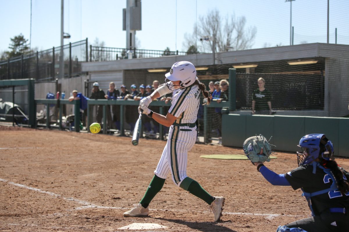 Woman in white and green baseball attire and a white helmet hits a yellow ball with a bat that flies forward, her back right foot off the ground.