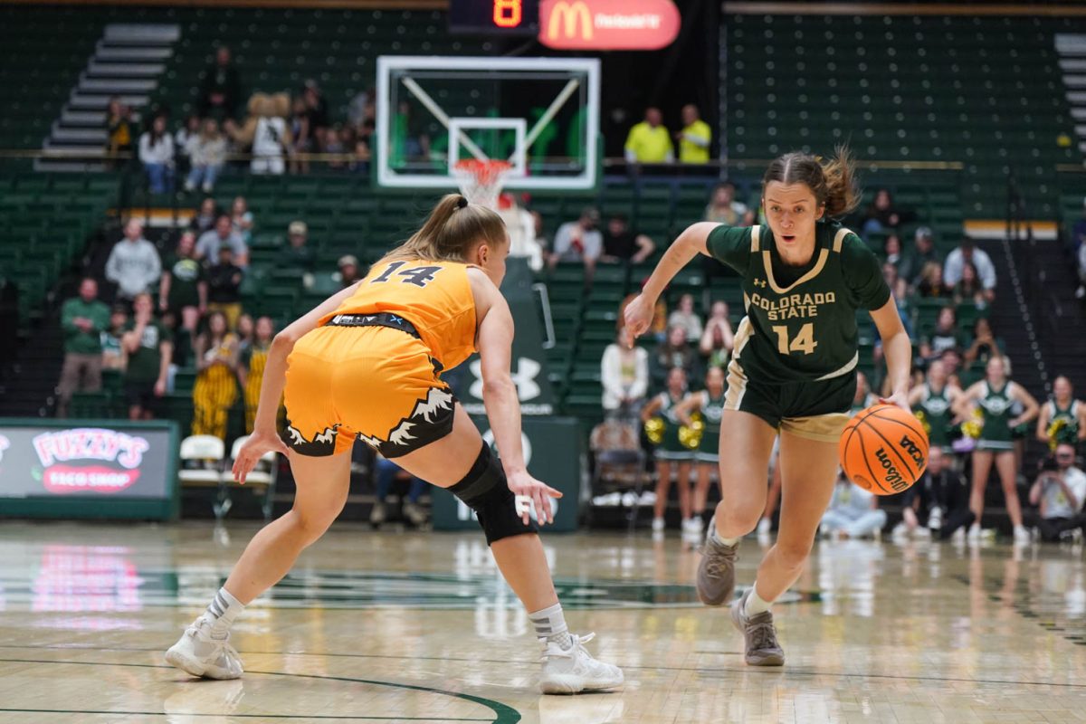 A woman in a green jersey holds an orange ball with one hand and runs past a woman in a yellow jersey.