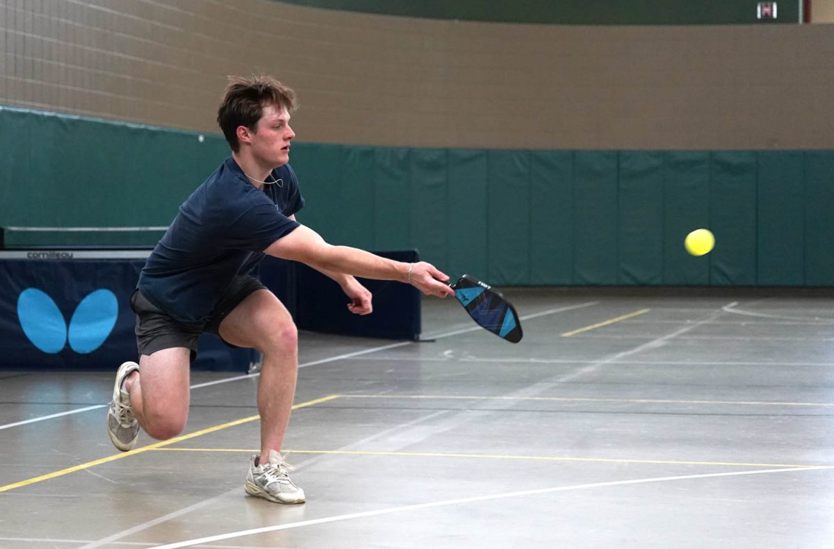 A man with a blue shirt hits a yellow ball with a pickleball racket.