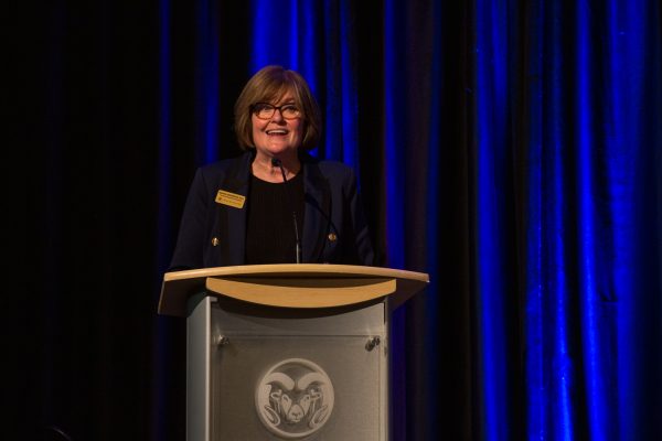 A woman stands on stage speaking behind a podium with a blue curtain in the back.