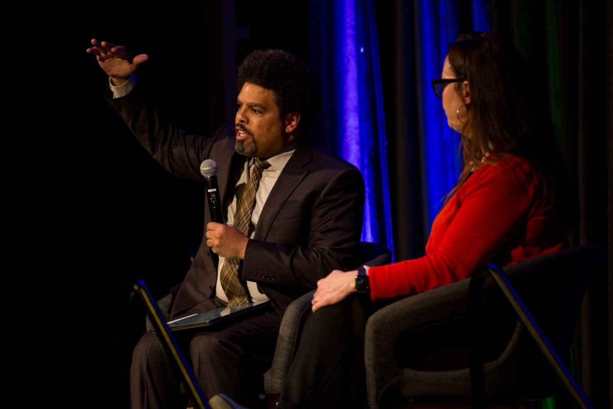 A man and woman sit on chairs on a stage together in front of a blue and green lit curtain.