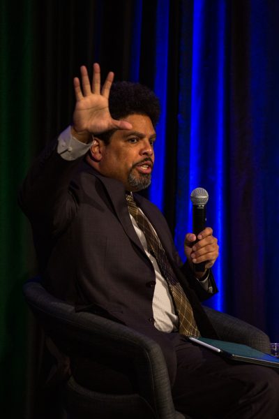 A man with a microphone sit on a chair on a stage in front of a blue and green lit curtain.