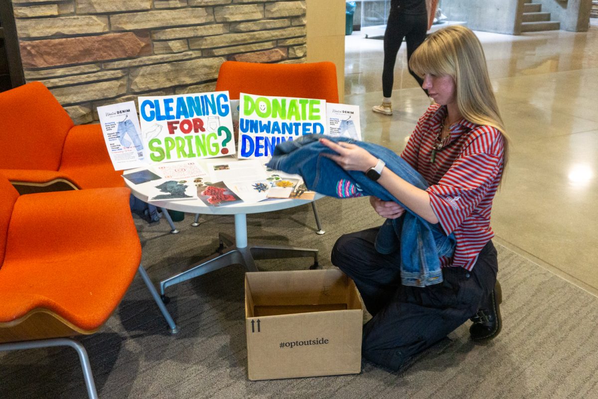 A college student kneels in front of the table with signs that say "Cleaning for Spring? Donate Unwanted Denim Here" in bold letters.