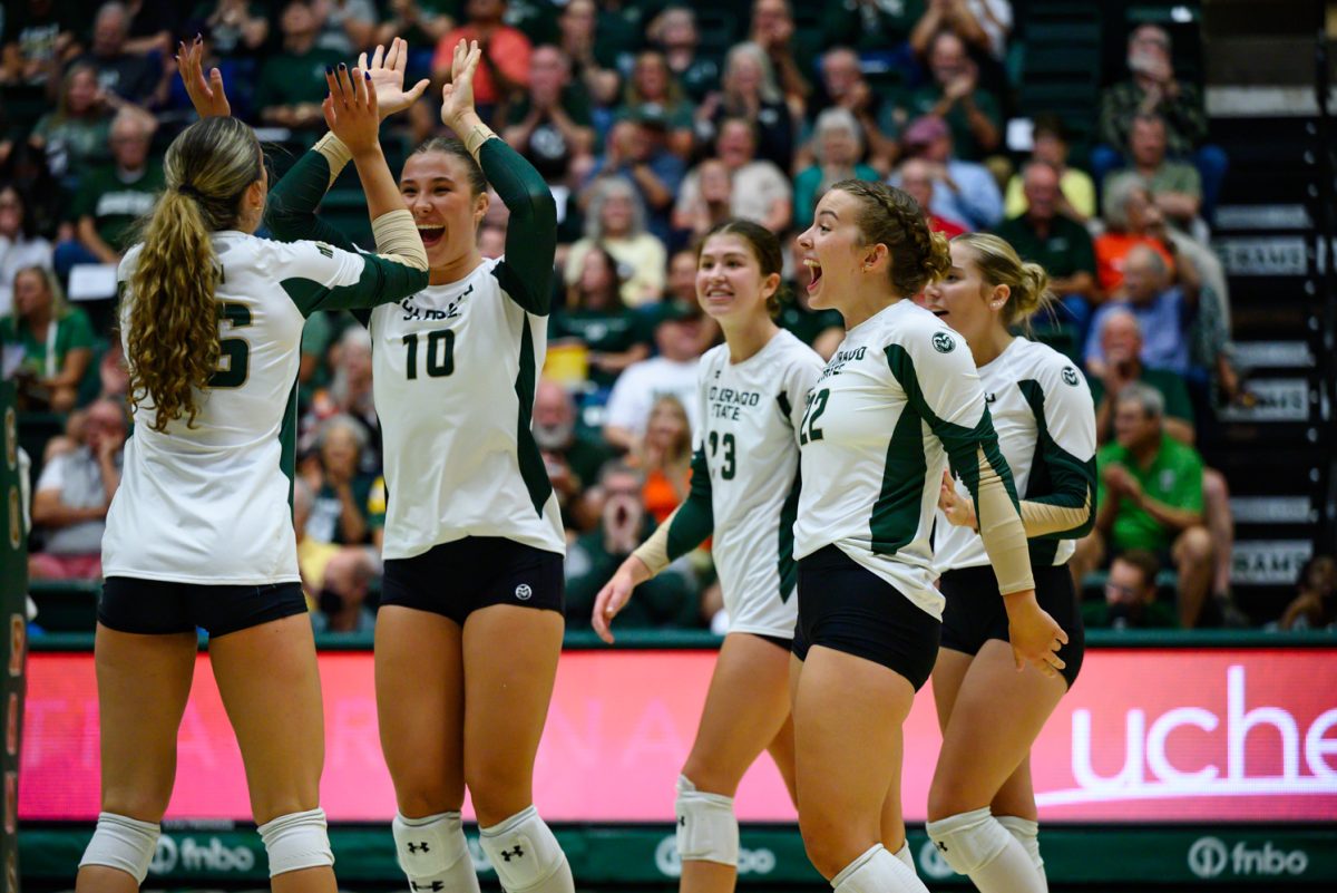 Two women's volleyball players in white and green uniforms smile and high five while three more players cheer and walk to them.