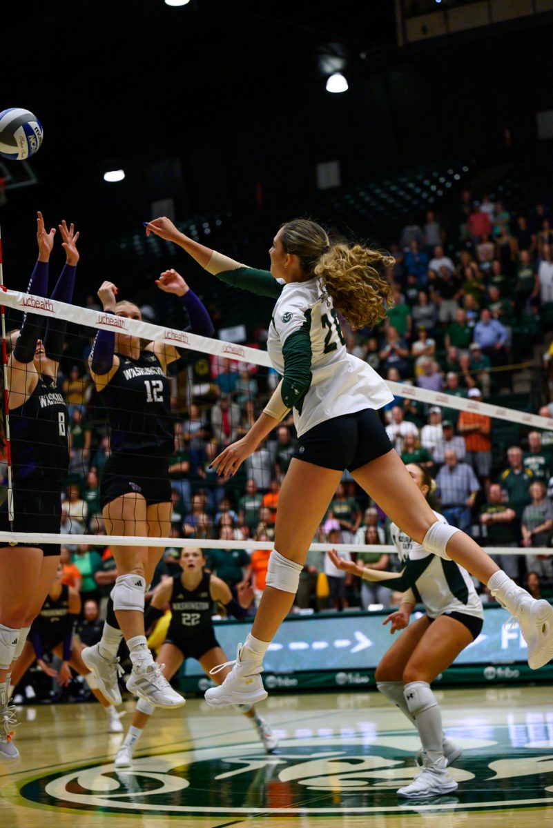 A women's volleyball player in a white and green jersey hits a volleyball over the net at two players in black and purple jerseys.