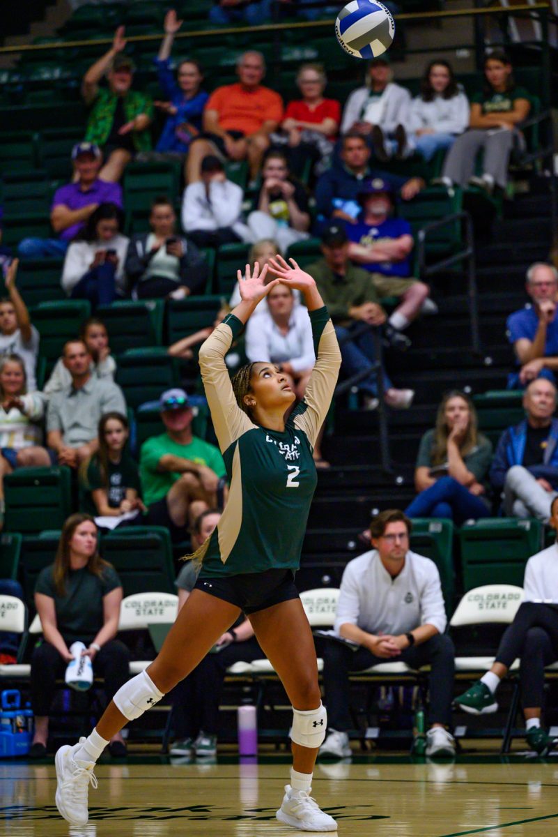A women's volleyball player in a green and gold uniform looks up at a volleyball in the air so she can set it.