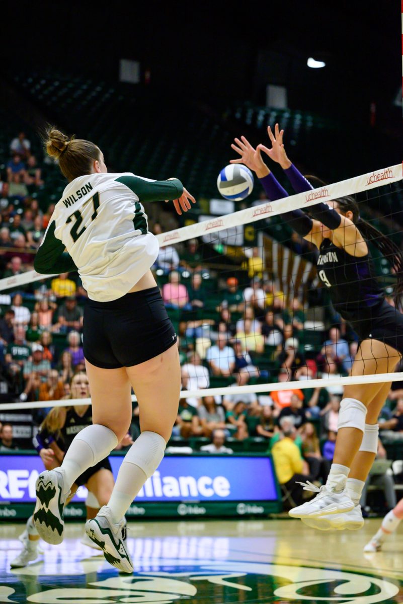 A volleyball player in a white and green uniform spikes a volleyball over the net at a player in a black in purple jersey.