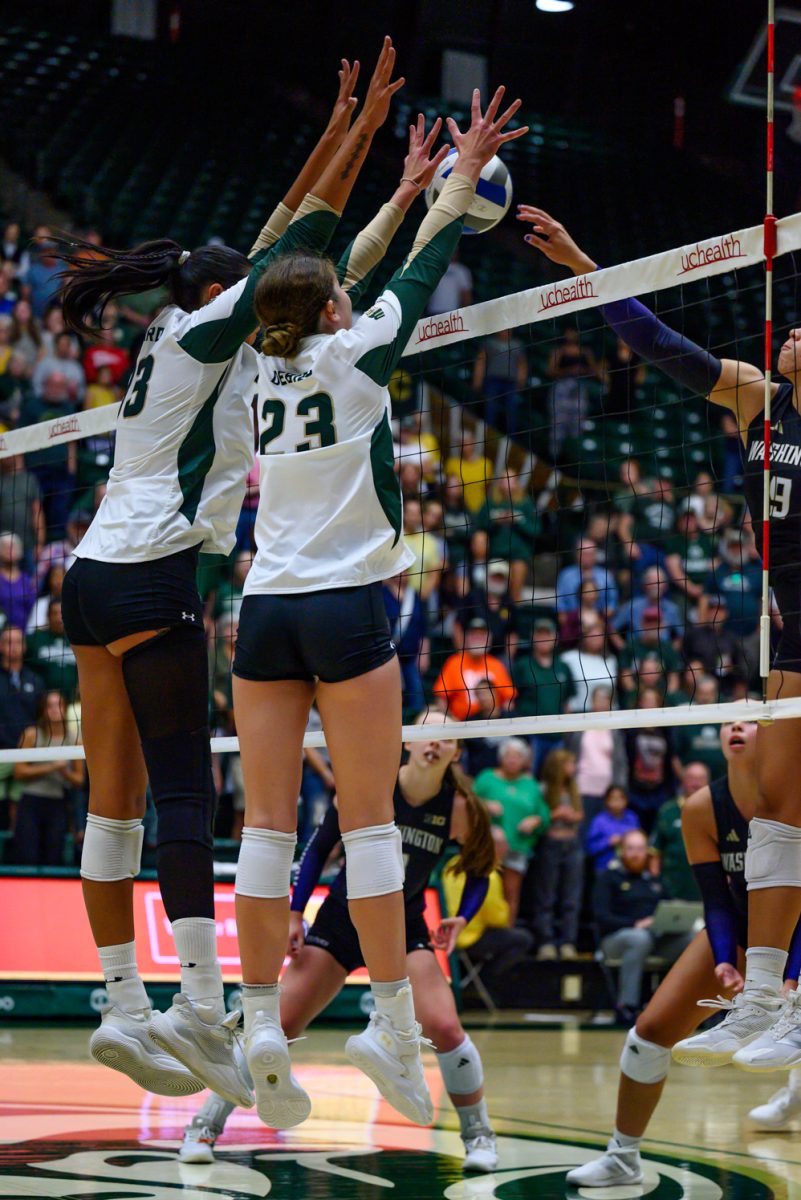 Two women's volleyball players in white and green jerseys block a volleyball from going over the net.