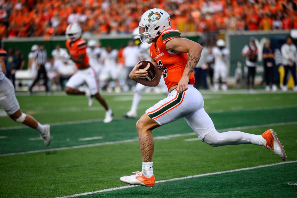 A football player in orange and white runs with a ball while players in all white pursue him.