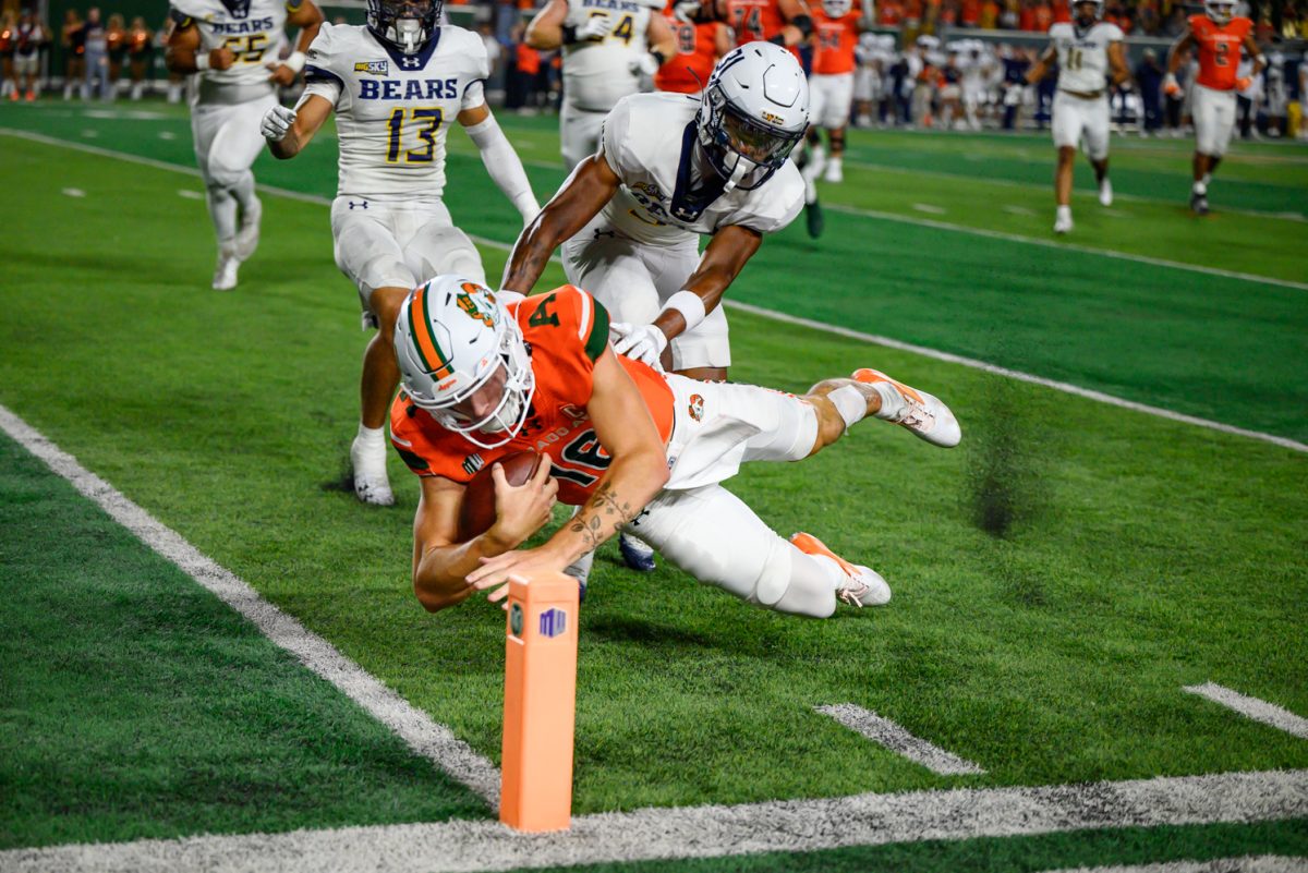 A football player in orange and white clutches a ball to his chest while diving into the end zone. A player in all white tries to grab him, but misses.