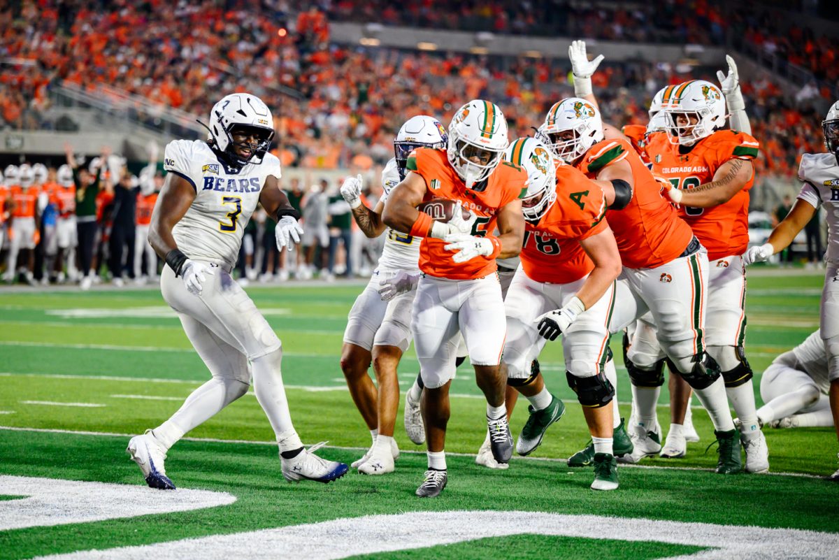 A football player in orange and white holds a ball and runs into an end zone with his teammates behind him. A player in all white runs alongside.