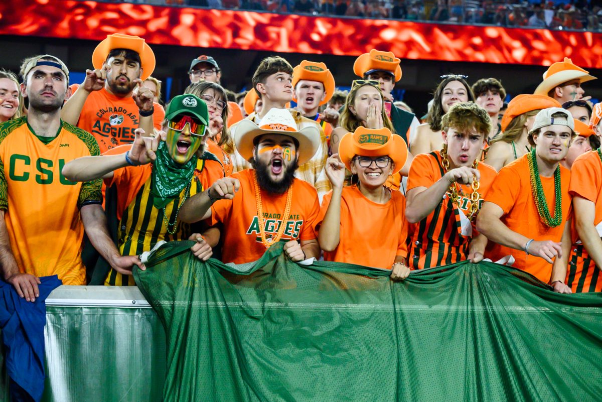 A crowd of college students in orange Aggies merch hold up a green flag and make the ram sign with their hands while cheering.