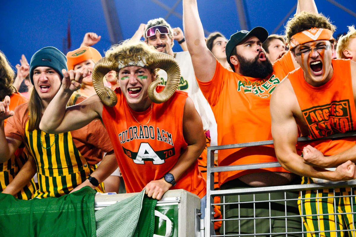 A crowd of college students yell and make the ram sign with their hands while cheering. They are in orange Aggie merch, with one student having a set of ram horns on his head.