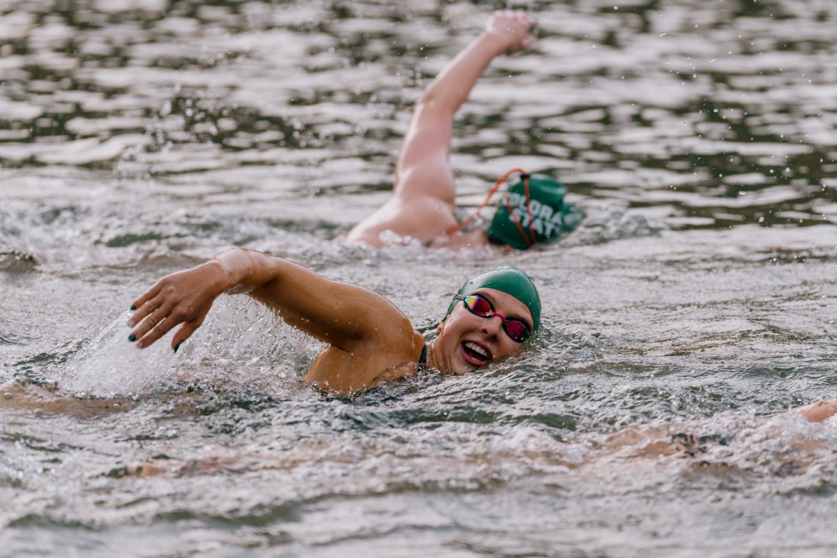 A swimmer in a green Colorado State cap pauses mid-stroke and lifts her head above the water to smile to the camera.