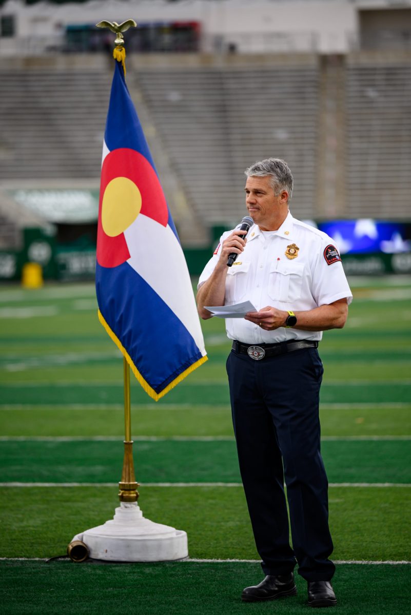 A man in uniform smiles and speaks into a microphone. Behind him sits a single Colorado flag.