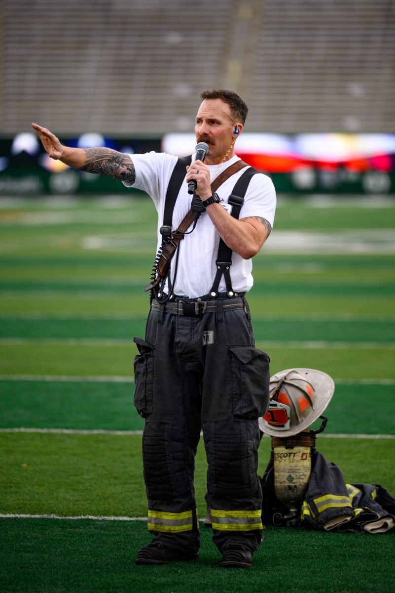 A mustached man in firefighter gear gestures off to the left and speaks into a microphone.