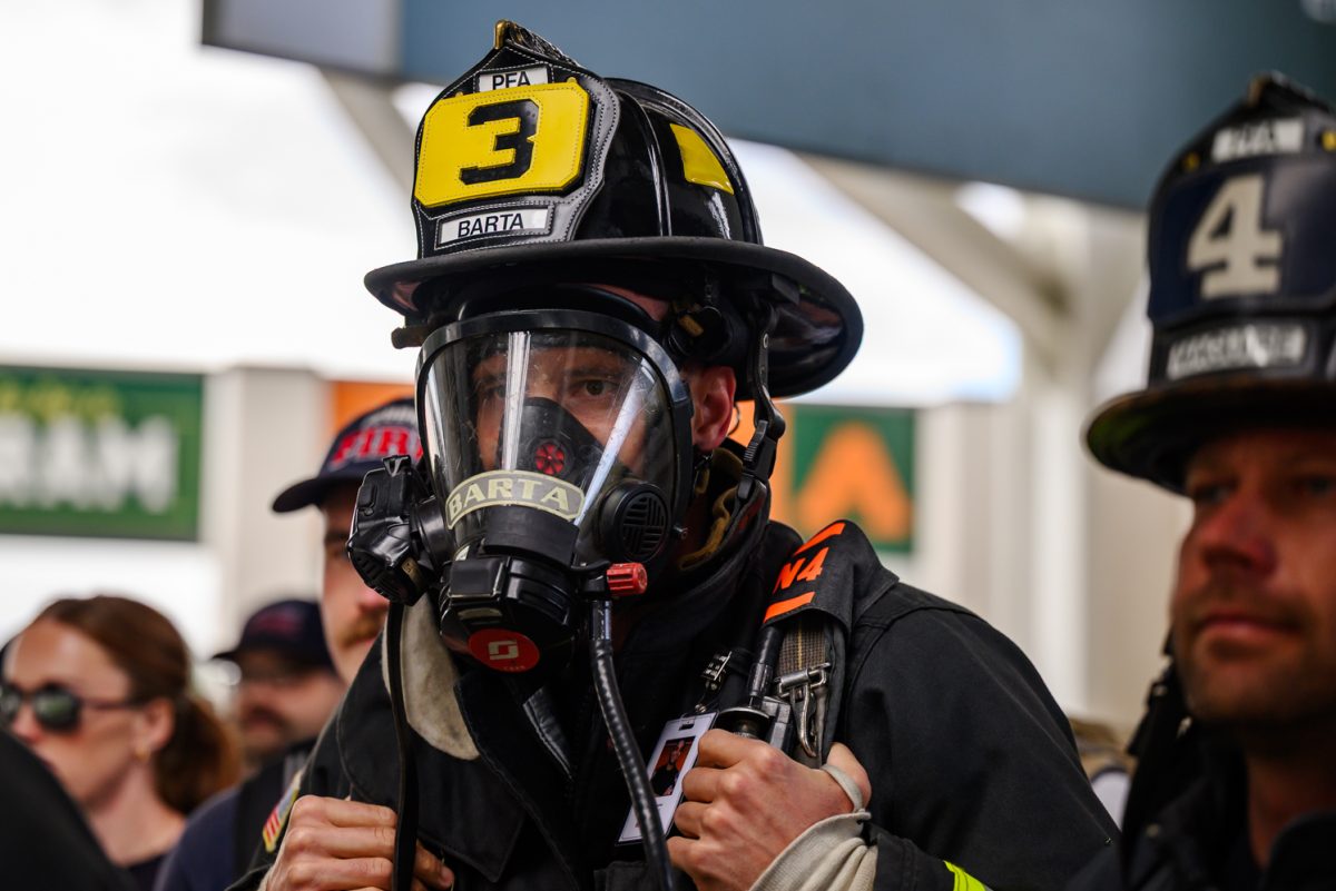 A male firefighter in full uniform clutches the straps of his backpack while waiting in line. His face is covered by a full respirator.