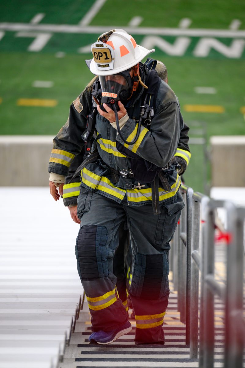A fully uniformed firefighter adjusts his full-face respirator while leading a line of people up a row of stairs.