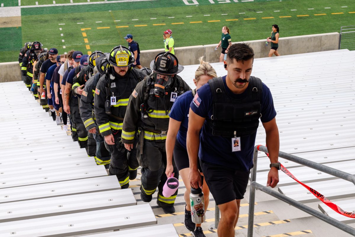 A line of people walks up a row of stairs. Half of the people are in full firefighter uniforms and gear, with others wearing EMS hats or American flags.