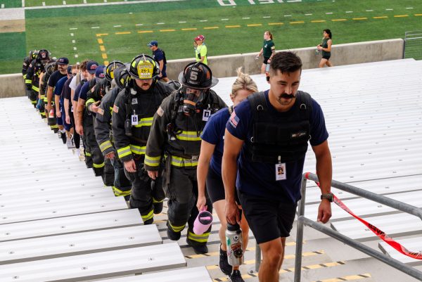 A line of people walks up a row of stairs. Half of the people are in full firefighter uniforms and gear, with others wearing EMS hats or American flags.
