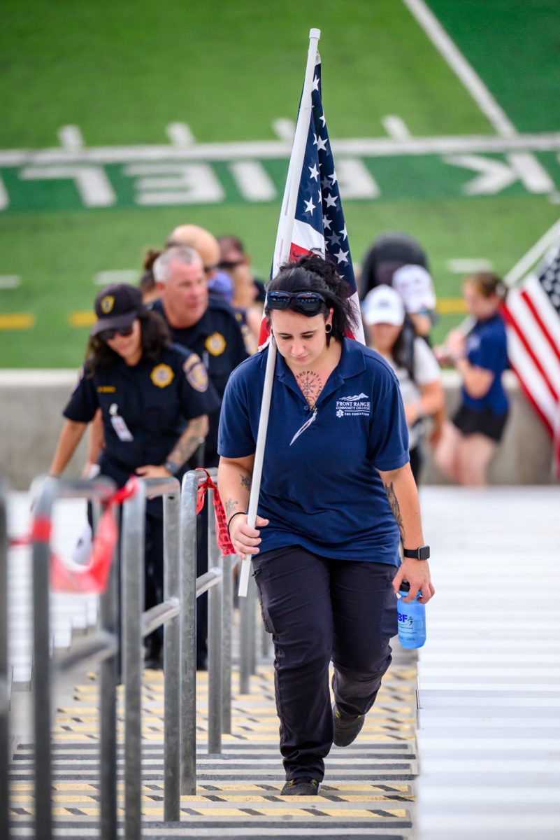 A woman in a blue Front Range Community College EMS Education shirt holds an American flag over her shoulder and a water bottle by her side while walking up stadium stairs.
