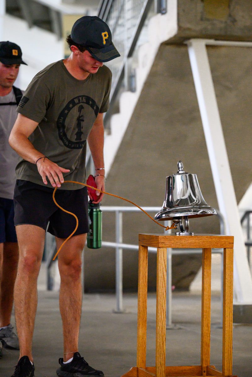 A man in a green shirt and baseball cap releases an orange rope after using it to ring a large silver bell.