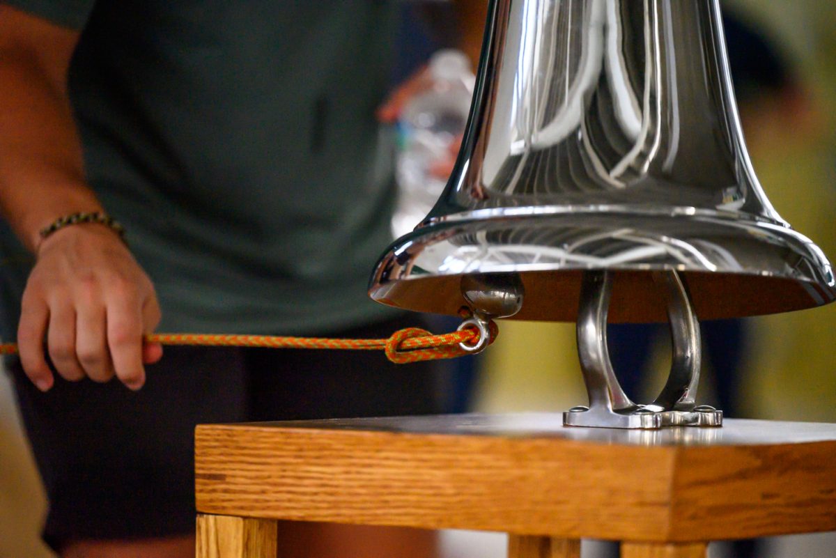 A large silver bell is rung by a hand pulling an orange rope.