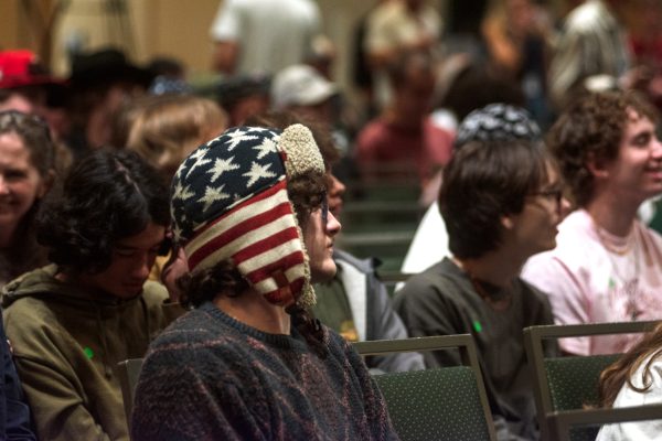 A young man is wearing a beanie decorated with an American flag themed design while sitting in an audience amongst others.