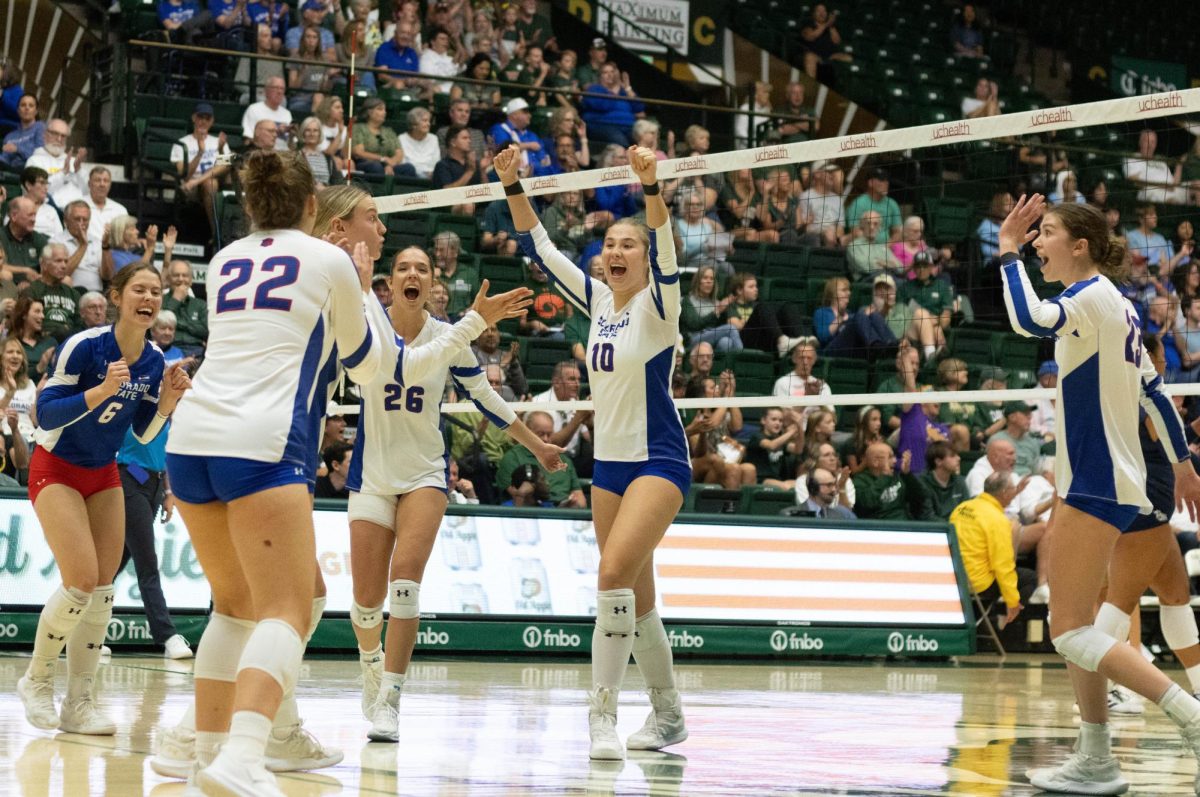 5 women wearing white jerseys and blue shorts and 1 women wearing a blue jersey and red shorts celebrating with their hands up. They have smiles on their faces.