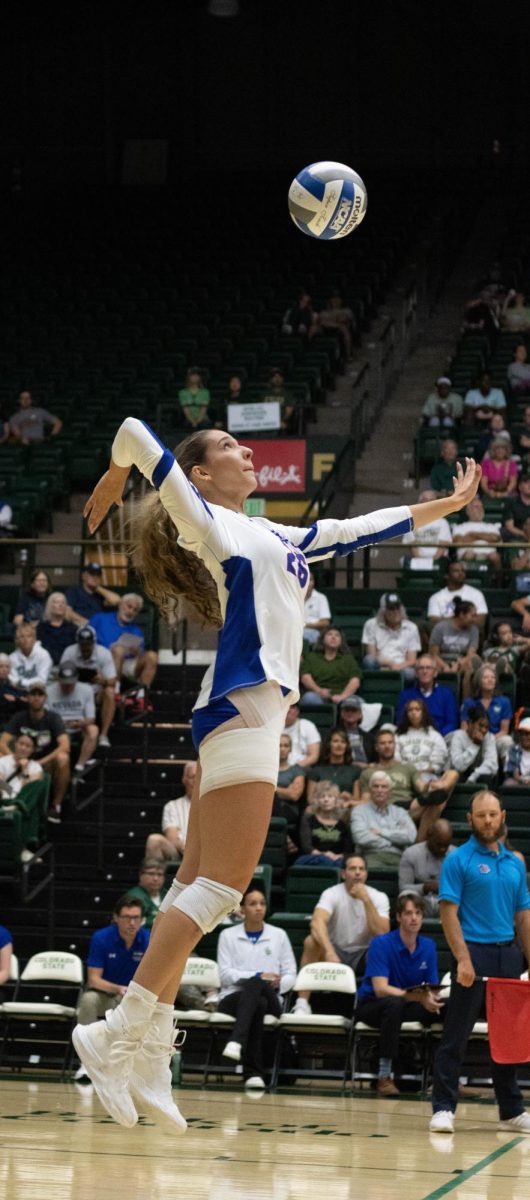 A women wearing a white jersey and blue shorts jumps up with her hand extended out at the ball above her head. She has bandages wrapped around her upper right leg.