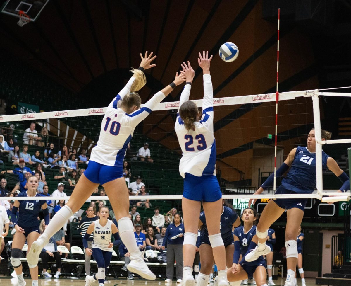 Two women wearing white jerseys and blue shorts jump above the net with both of their hands extended over their head at the ball.