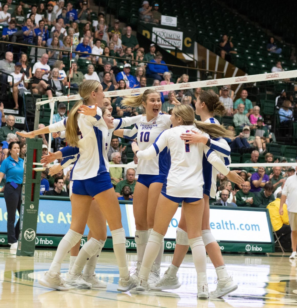 5 women wearing white jerseys with blue shorts and 1 women wearing a blue jersey and red shorts hugging each other with smiles on their faces.