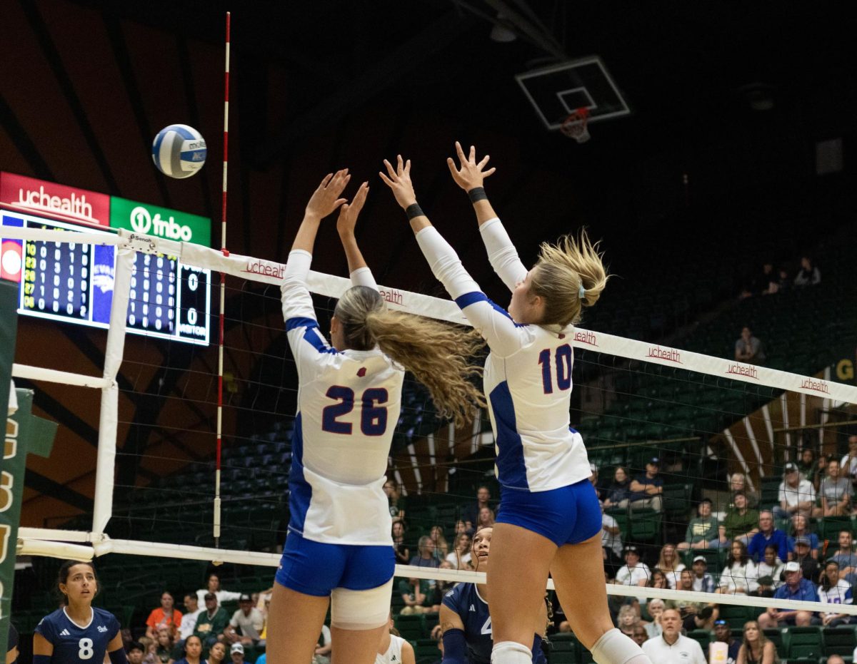 Two women in white jerseys and blue shorts jump with both of their hands extended at the ball over the net.