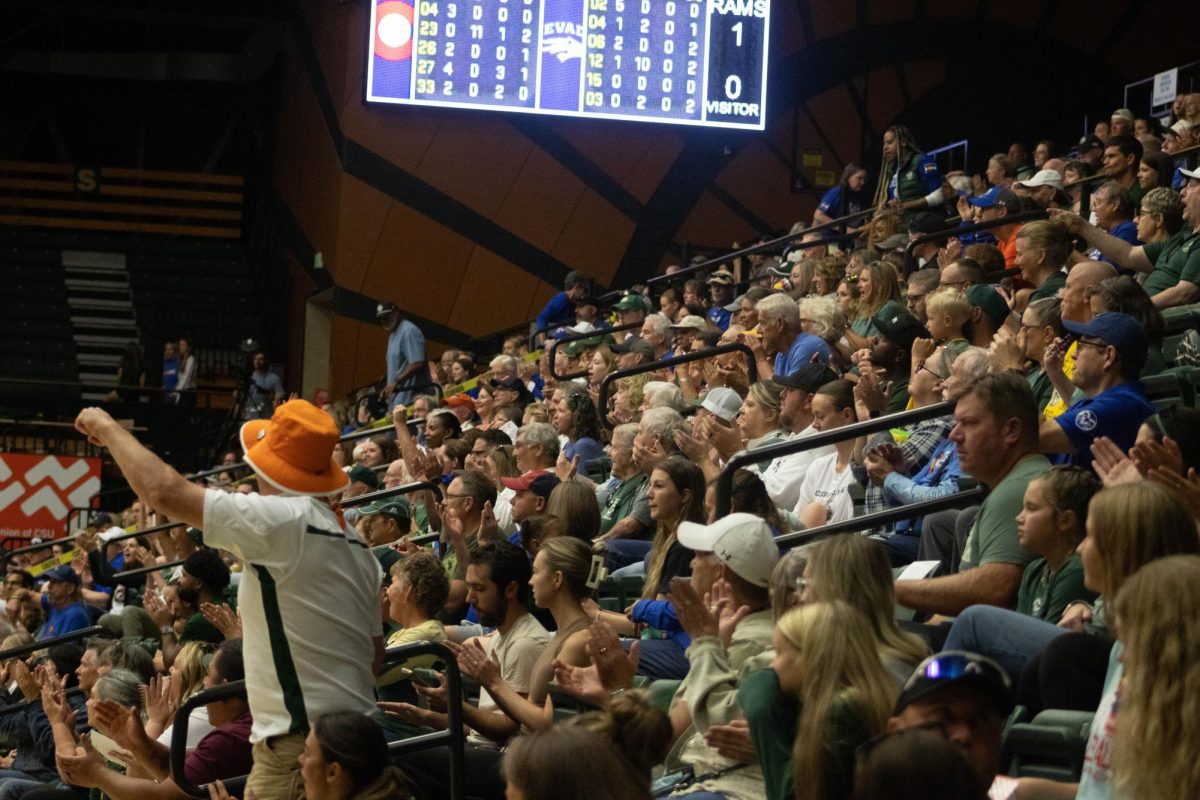 A crowd of people clapping their hands together. One man is standing wearing an orange bucket hat.
