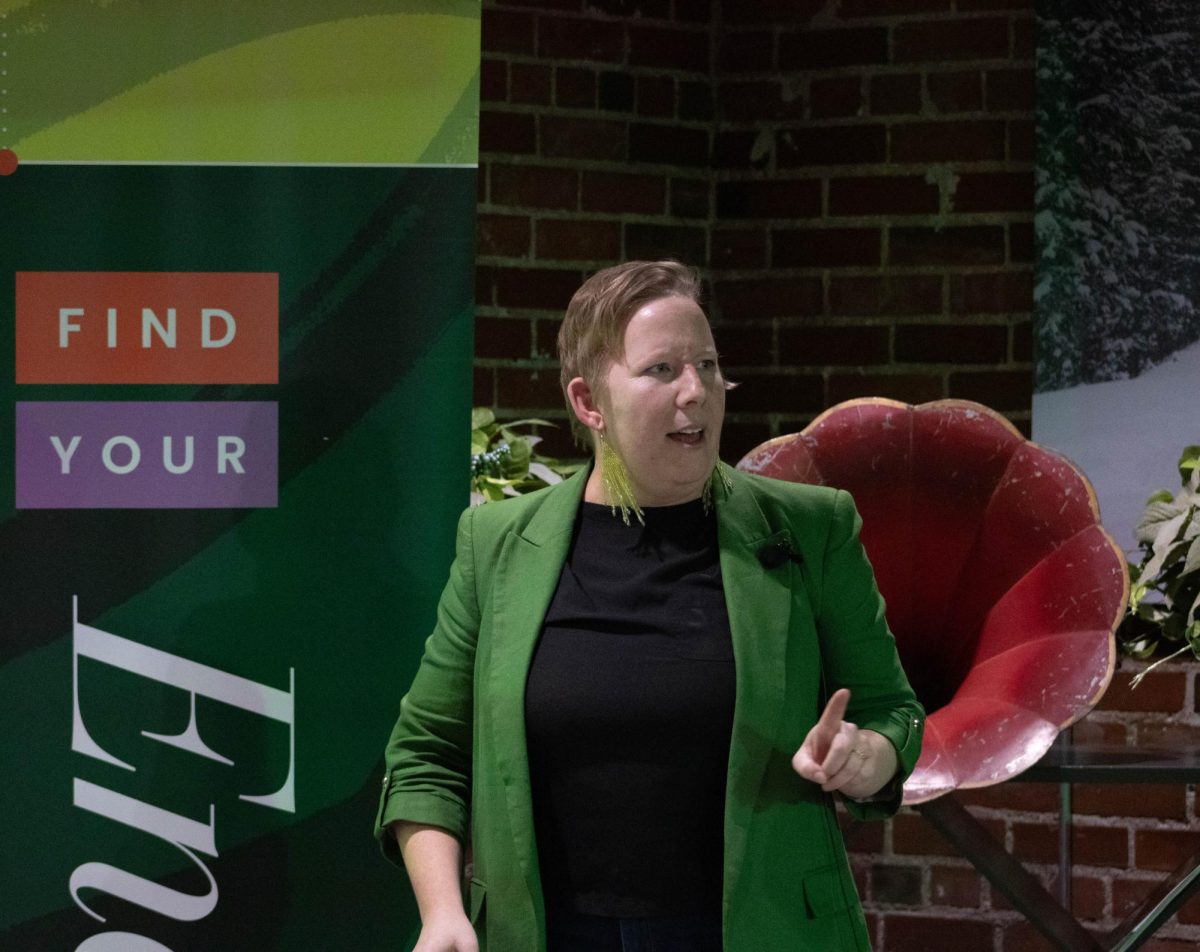 A women wearing a black shirt, green blazer and green reflective earrings talks with her hand pointing to the audience.