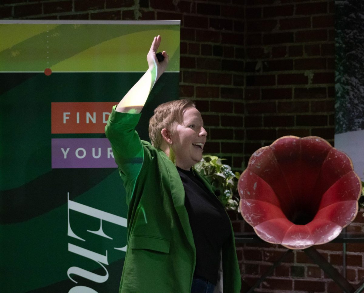 A women in a black shirt, green blazer, and green reflective earrings talks with her hand above her head.