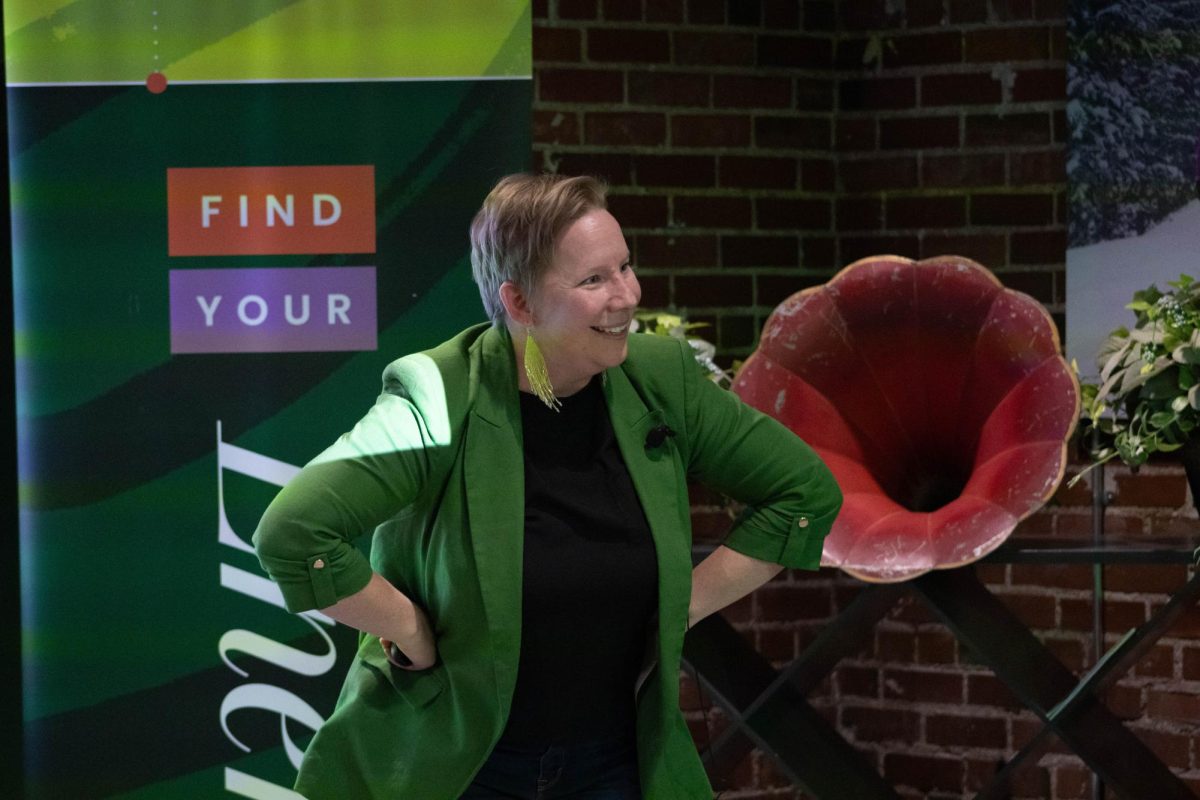 A women wearing a black shirt, green blazer and green reflective earrings with her hands at her hips, leaning slightly forward with a smile on her face.